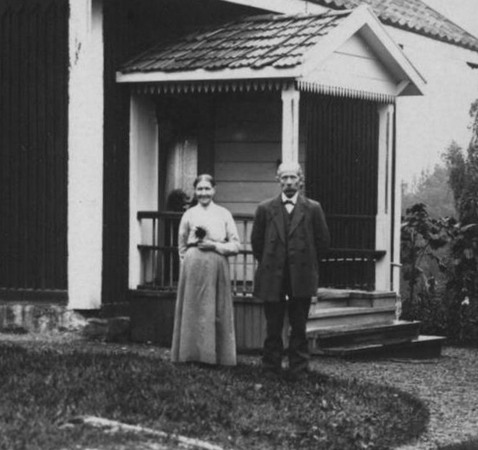 En man och en kvinna står finklädda framför ett litet hus med snickarglädje.