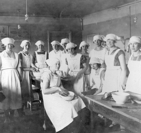 12 unga flickor iklädda vita förkläden och huvudduk i ett kök med bunkar och slevar.