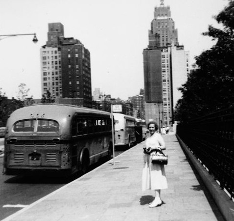Kvinna på trottoaren, buss på gatan och två skyskrapor i fonden.