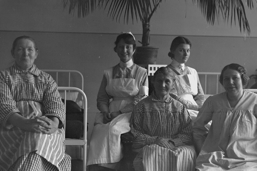 Tre kvinnliga patienter och två kvinnliga sköterskor framför sängarna i sovsalen. En palm i bakgrunden