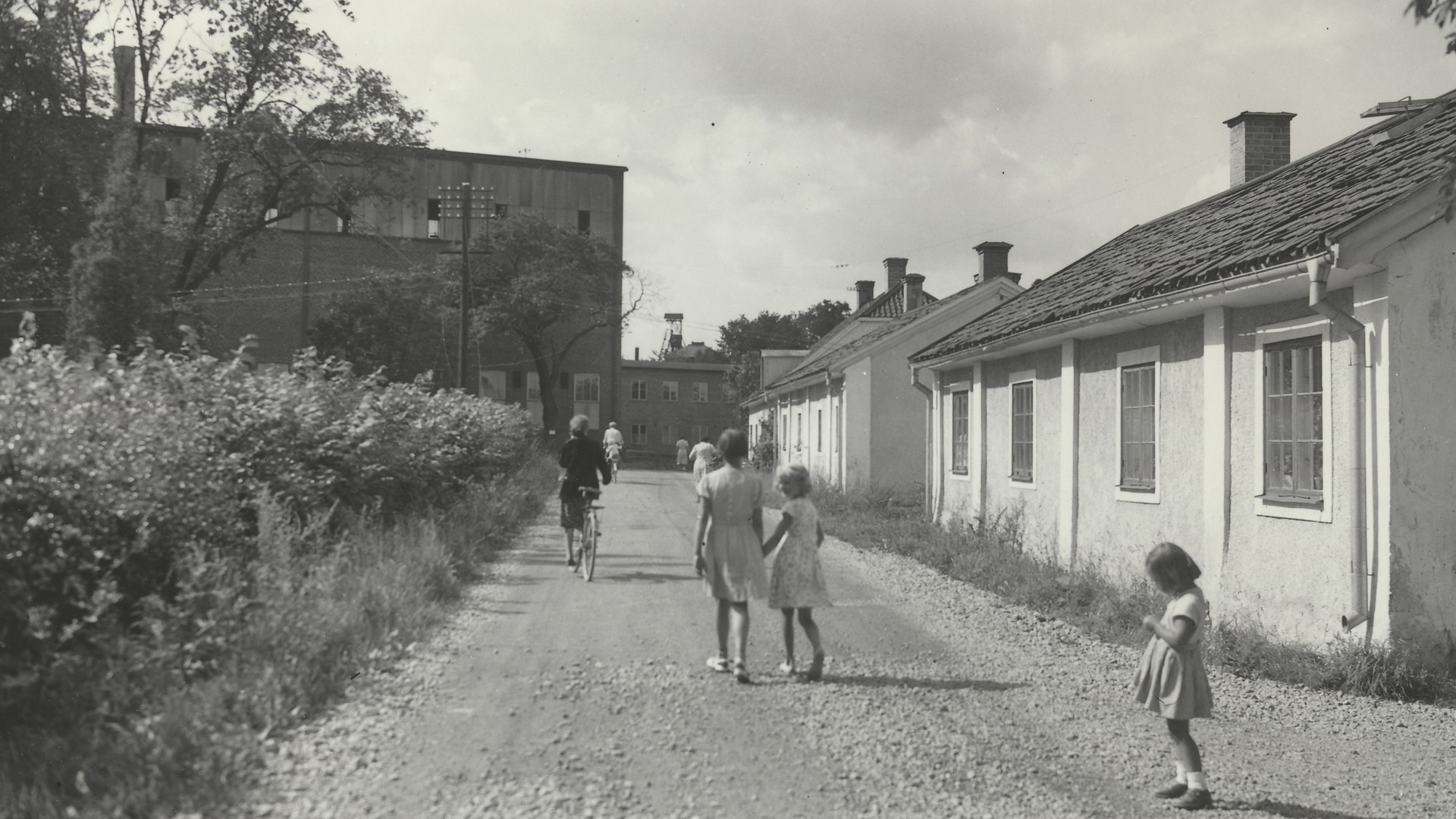 svartvit bild med barn på grusväg, putsat hus till höger och fabriksbyggnad rakt fram.