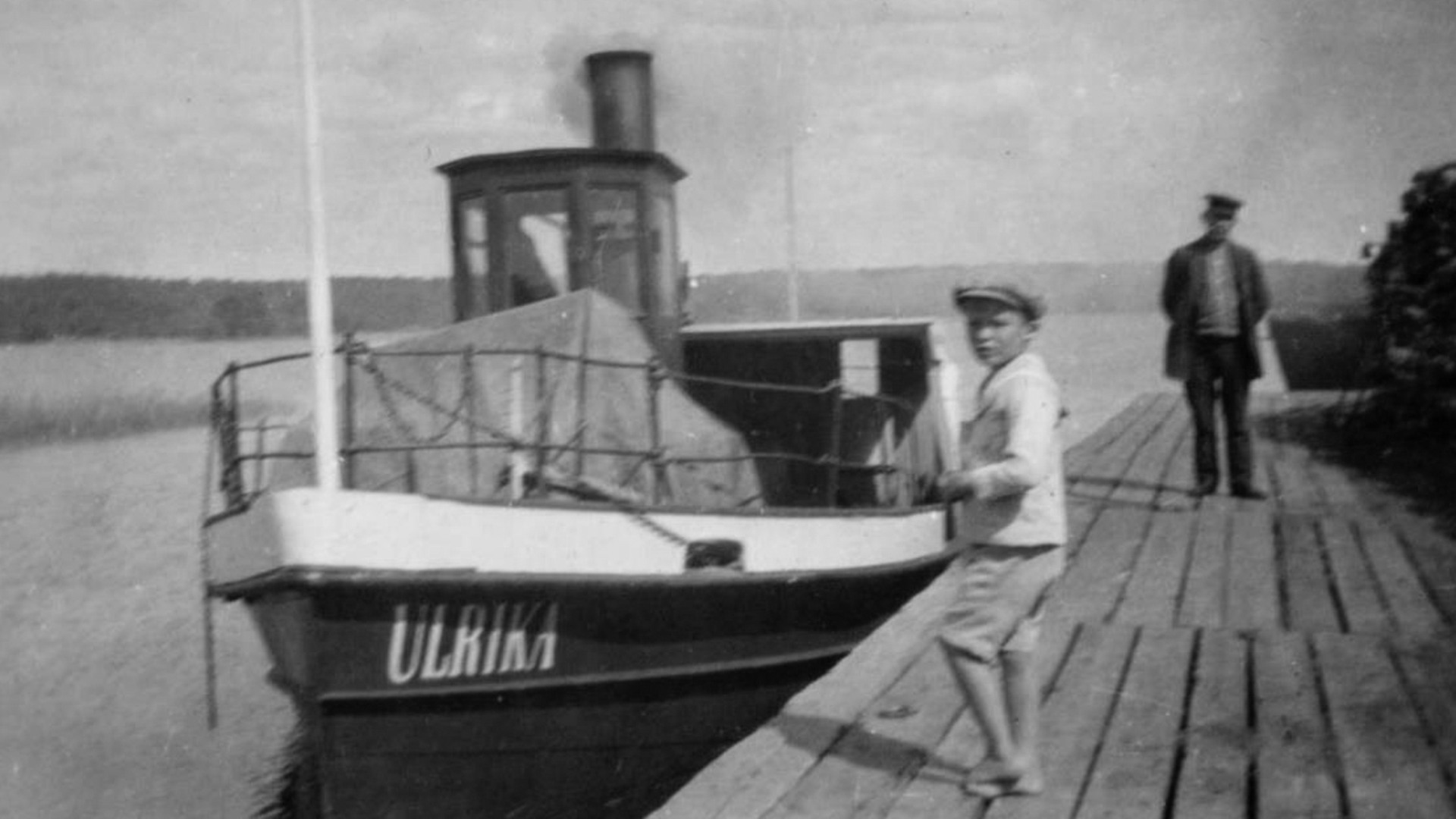 Ångbåten Ulrika ligger invid en brygga. Närmast kameran på bryggan står en pojke i keps och lite längre bort en man.