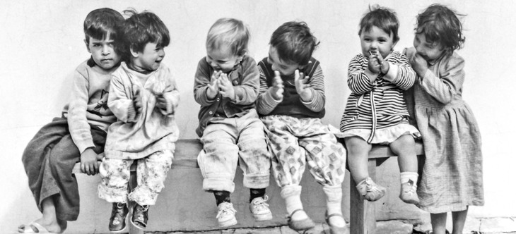 Sex små barn sitter bredvid varandra på en träbänk, ler åt varandra och klappar händerna