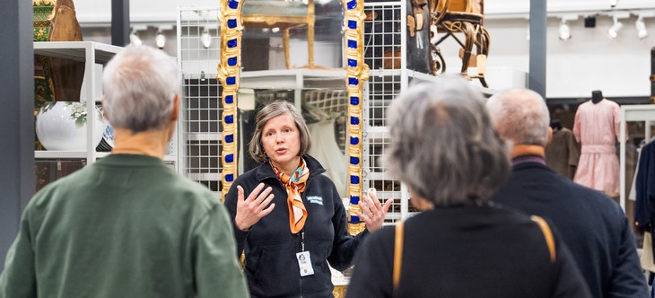 En grupp människor - barn och vuxna - går i ett museums magasin och beundrar föremål. 