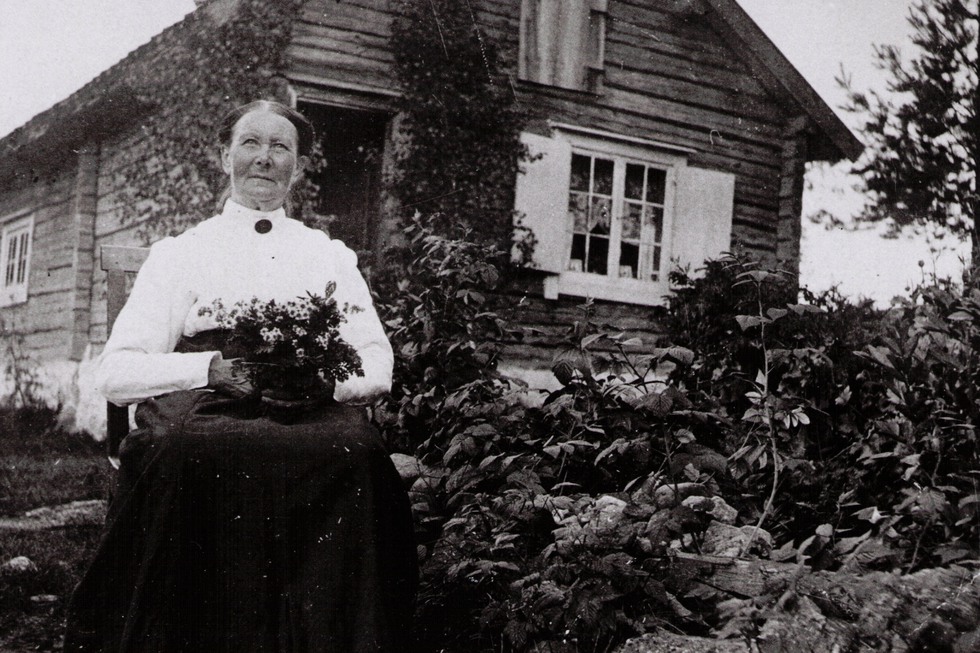 Svartvitt foto, äldre kvinna framför stuga.