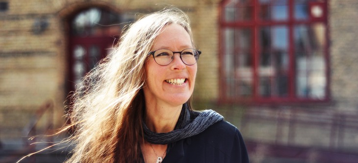 En blond leende kvinna med glasögon, i svart topp och mörk scarf.