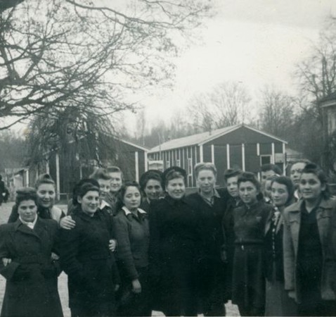Gruppfoto, kvinnor som överlevt förintelsen. Bostadsbaracker i bakgrunden.