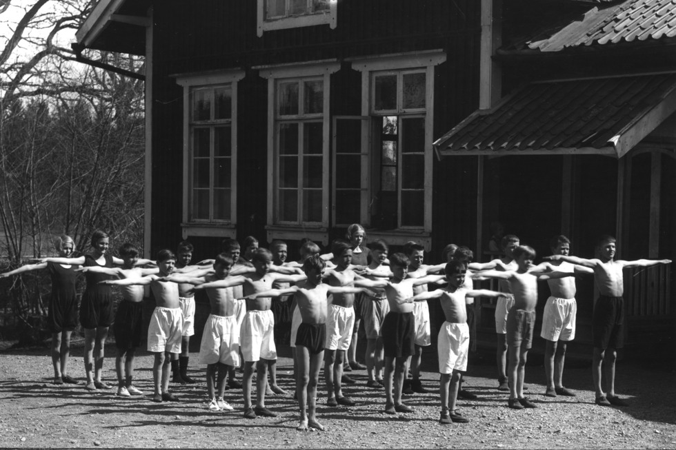 Svartvitt foto gymnastik utomhus, stor grupp håller armarna rakt utsträckta.