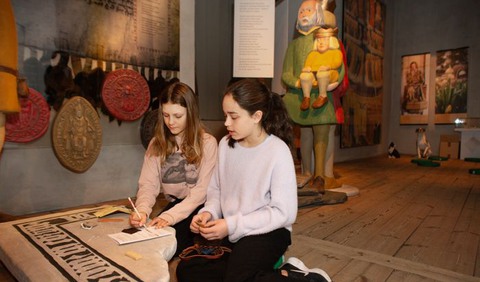 Två flickor - den ena med papper och penna - sitter på golvet framför en stor sten med tecken.