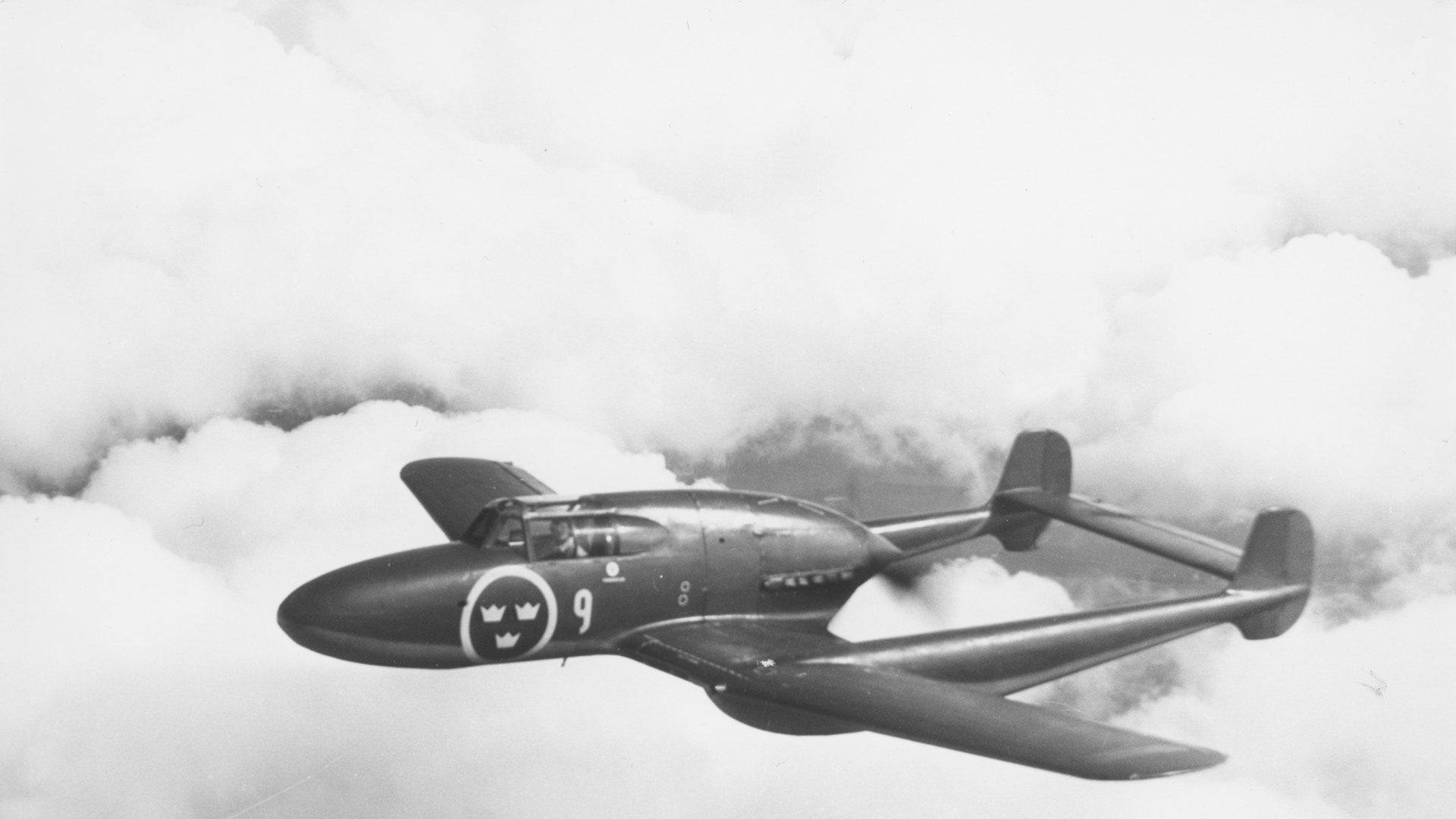 Motiv: Flygplan J 21A tillhörande F 9 Göta flygflottilj i luften med moln som fond. Vy snett ovanifrån. Tillhör Flygvapenmuseums bildarkiv.