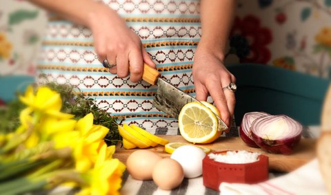 Kvinnohänder skär upp en citron med en kniv med träskaft, kvinnan bär ett förkläde, och på bordet ligger påskliljor, ägg, salt och halva rödlökar.