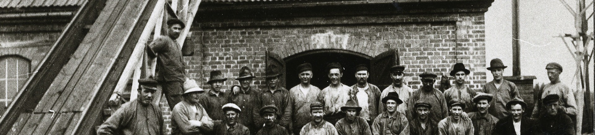 En grupp med män, alla arbetare, står uppställda framför ingången till industribyggnaden