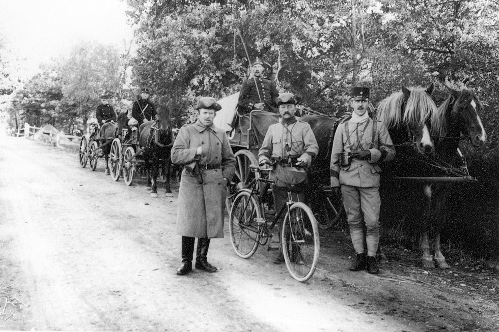 Några soldater på en väg, en har en cykel med sig och i bakgrunden står två hästar