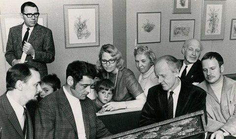 Ett svart-vitt foto på flera människor som sitter i en utställningssal och tittar tillsammans på en målning.