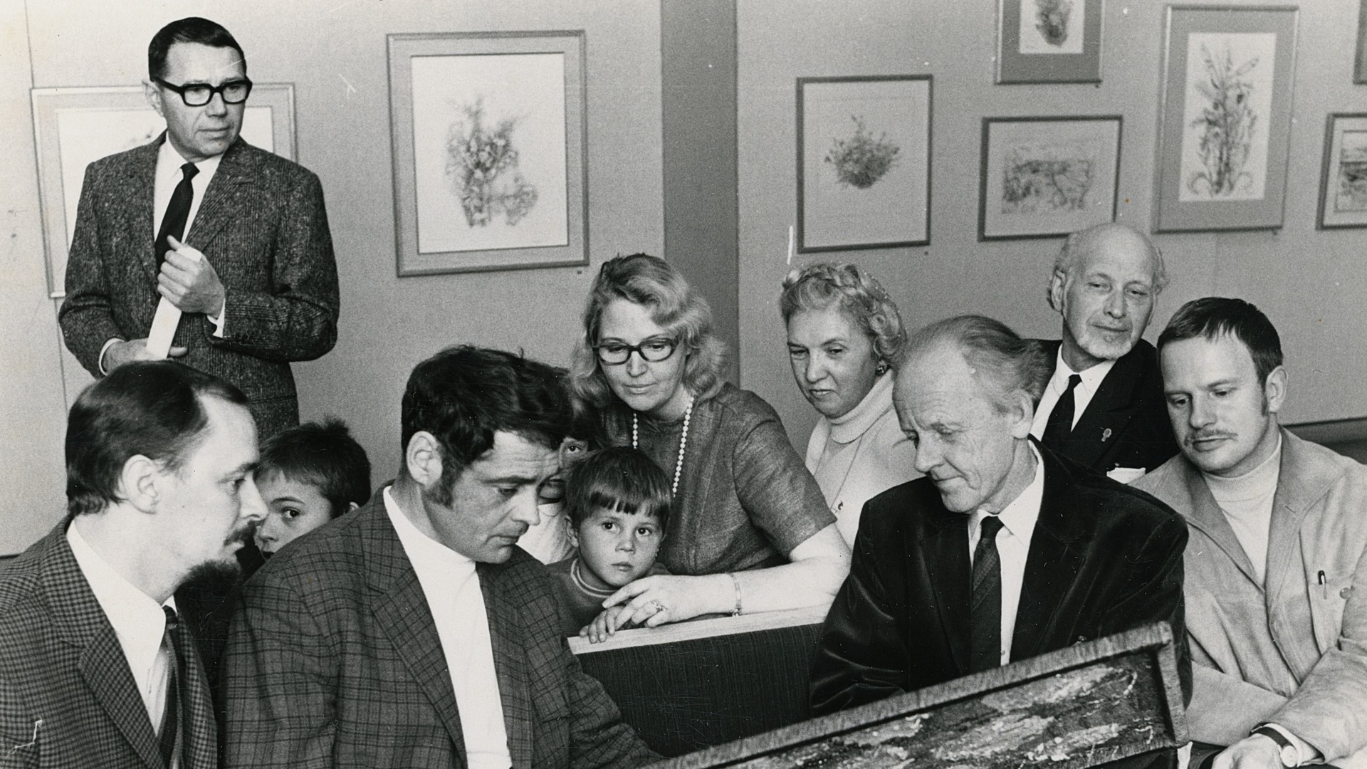 Ett svart-vitt foto på flera människor som sitter tillsammans i en utställningssal och tittar på en målning.