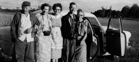 Fem personer framför en bil med öppna dörrar på ett fält.