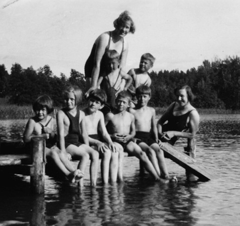 Två kvinnor och sju barn på en brygga. BArnen har fötterna i vattnet. I bakgrunden skog.