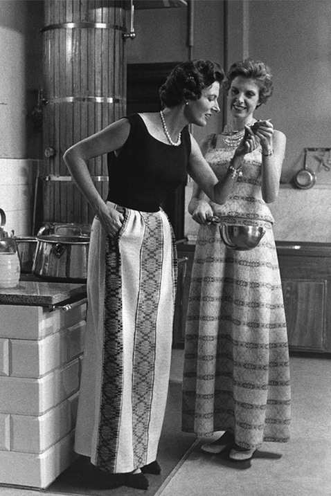 Två unga damer står i ett kök, iklädda lång kjol resp. lång klänning. Den ena kvinnan håller i en kastrull, medan den andra provsmakar från en sked. 