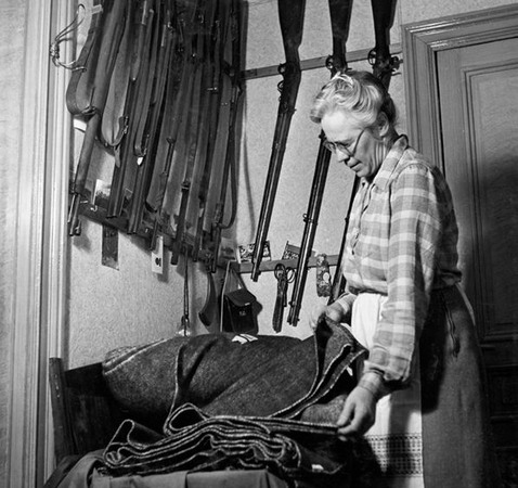 En äldre kvinna står bredvid en hög med hopvikta tyger och plockar upp det översta tyget. På vägger bakom henne hänger inte mindre än 10 gevär!