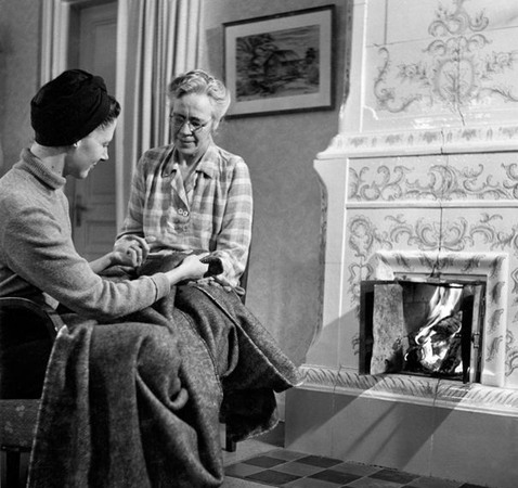 En äldre och en yngre kvinna sitter invid en kakelugn, med ett stort tyg i knät. De samtalar med varandra och tittar båda på tyget.