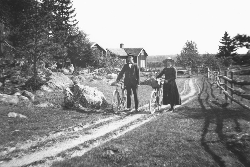 Man och kvinna med cyklar gåendes på landsväg, hus i bakgrunden.