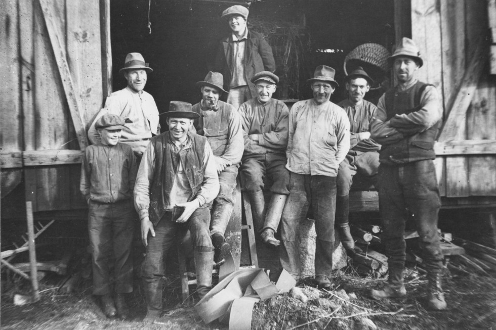 Svartvit bild med ett arbetslag bestående av män från barn till vuxen ålder, framför en ladugårdbyggnad.