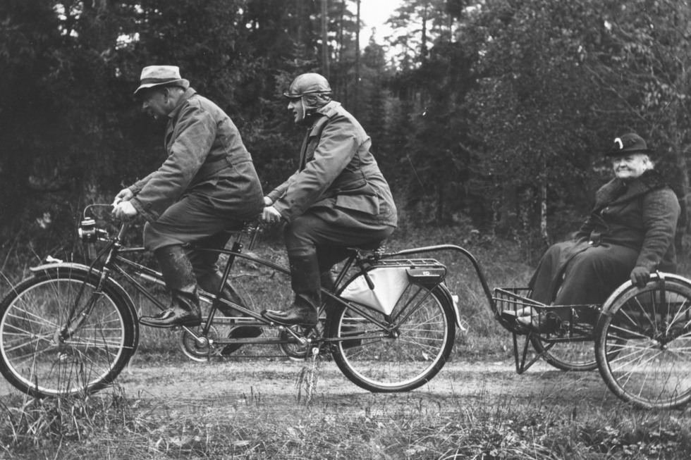 Två män på en tandemcykel, en kvinna på ett släp efter cykeln.