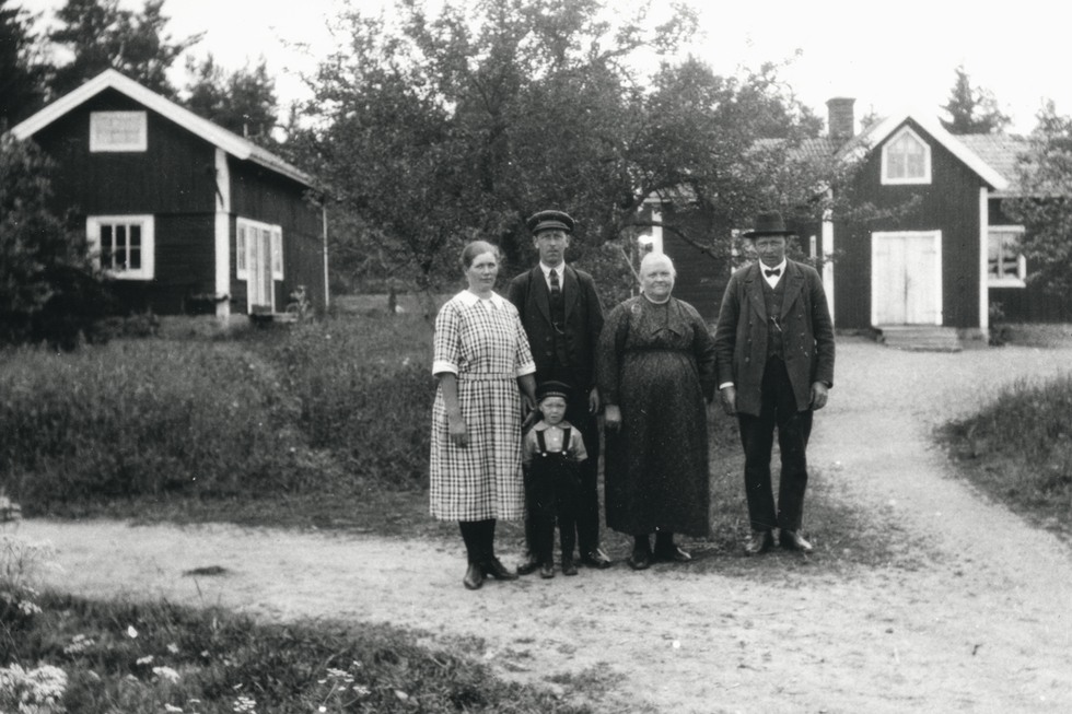 Svartvit bild med fyra vuxna och ett barn står uppställda på gårdsplanen, två hus i bakgrunden. 