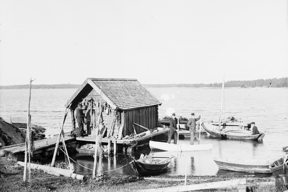 En brygga leder ut till ett litet trähus i havet, en man står vid huset och i en båt jämte står två till män.