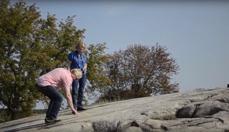 Två personer står på en klipphäll med hällristningar och pekar på ristningarna.
