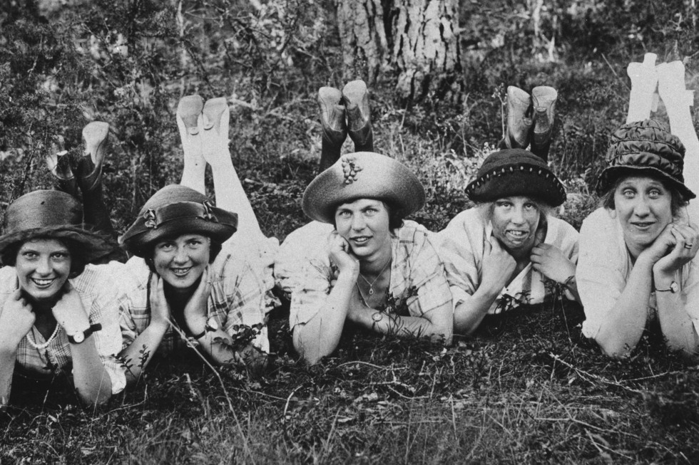 Svartvitt gruppfoto, fem unga kvinnor med hatt ligger i gräset.
